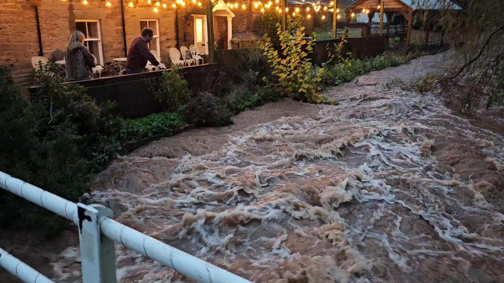 A very high river with choppy water runs alongside the pub while two people watch on. 