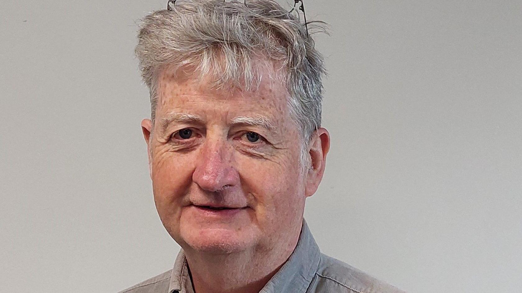 A man with grey hair and wire-framed glasses on his head. He is wearing a greay collared shirt, and standing against a plain light background in a council publicity shot.