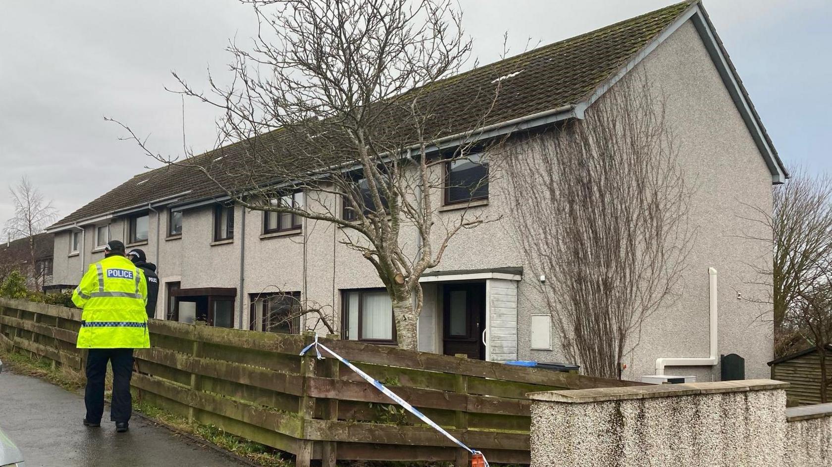 A semi-detached house in the Borders with a grey exterior and darker grey roof with two policemen standing outside and police tape across the driveway