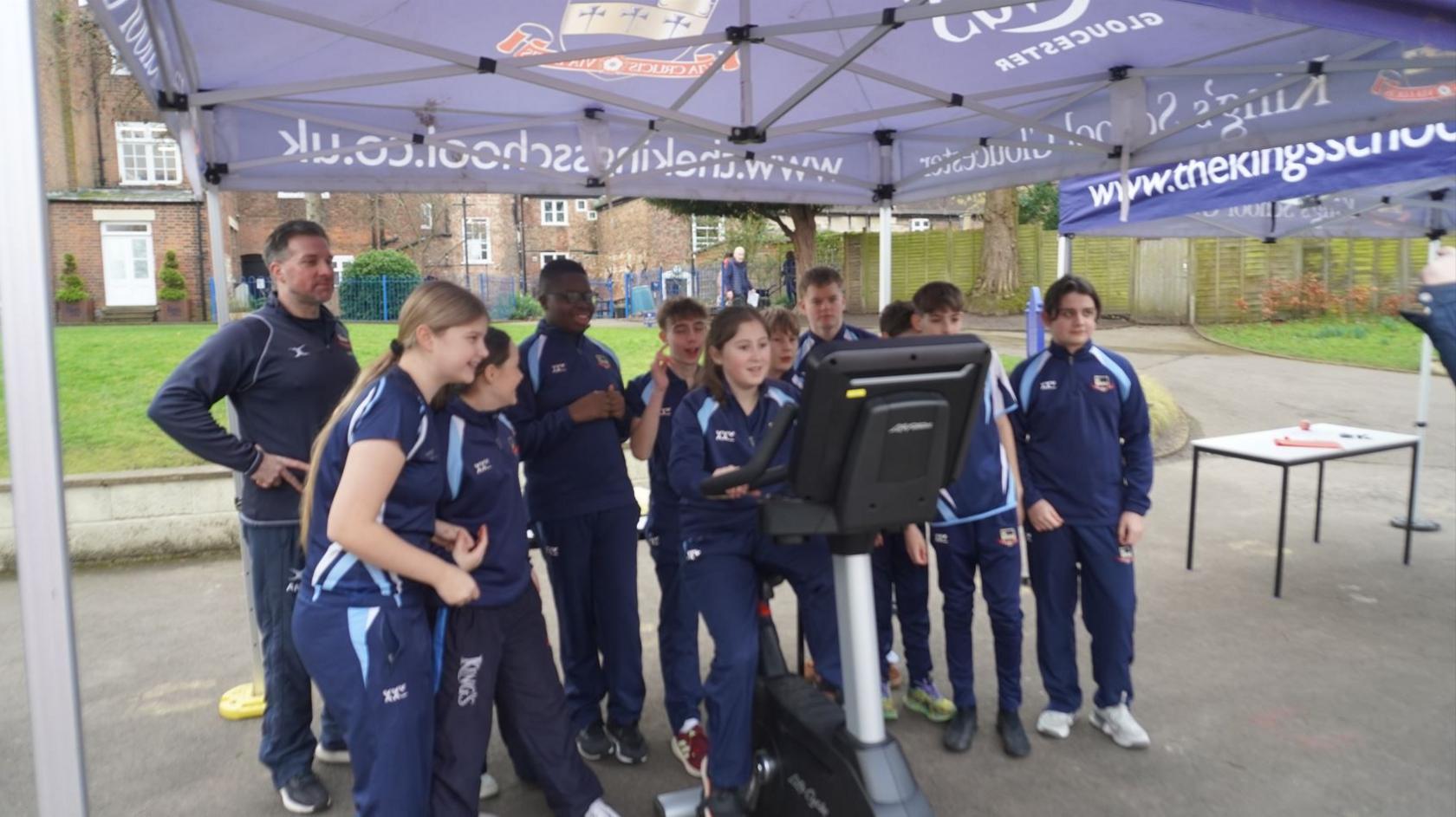 A group of pupils at The King's School in Gloucester, wearing their blue P.E kit uniform. There is a girl sitting on a static bike underneath a blue cabana, while the other children surround her and cheer her on. 