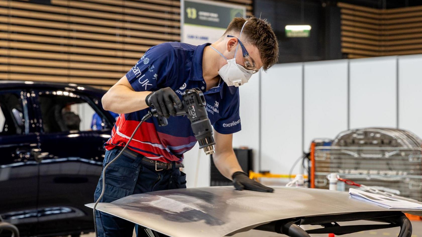 A man dressed in a navy and red Team UK branded polo shirt working on a car panel. He is wearing a mask and safety glasses. In the background is a black car.