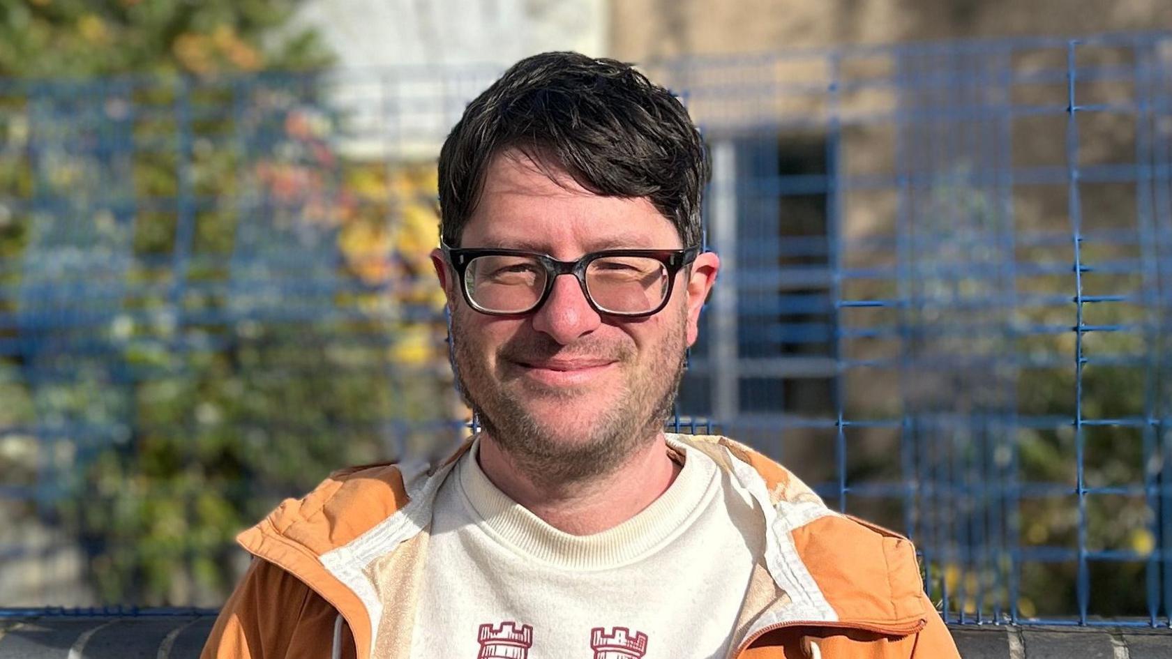 A person is seated outdoors on a bench, wearing an orange jacket over a light-coloured sweatshirt that has a design featuring two castle-like symbols. Behind the person, there is a blue wire fence and blurred greenery.