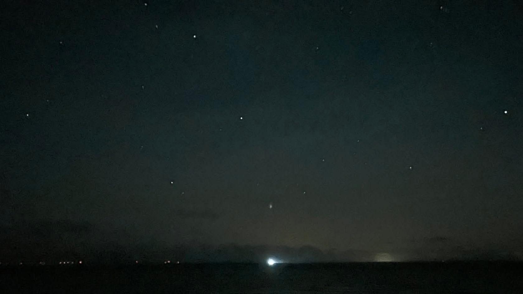 A dark stretch of sky with various bright white dots scattered across it. There is a light on the horizon.