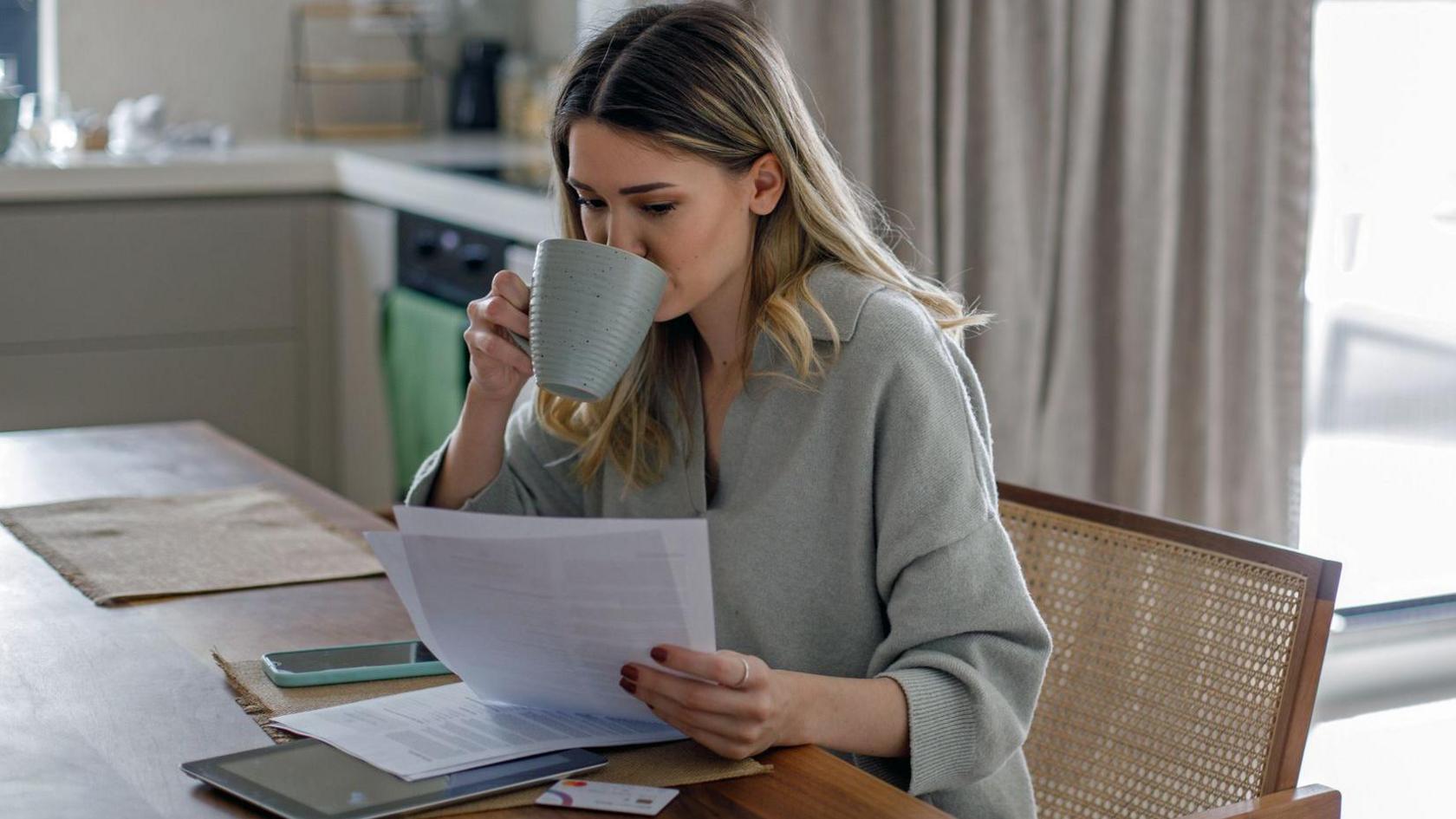 A woman sits in a chair at home looking at paperwork and drinking a cup of tea