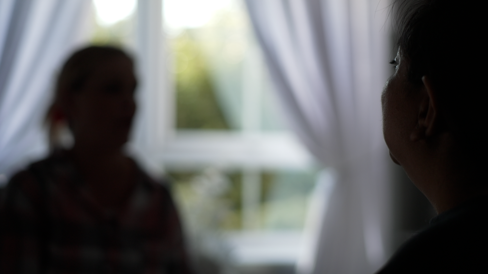 A blurred and blacked out image of a woman sitting in front of a window. The side profile of a second woman's face, taken from behind, can be seen in close-up to the right of the image.