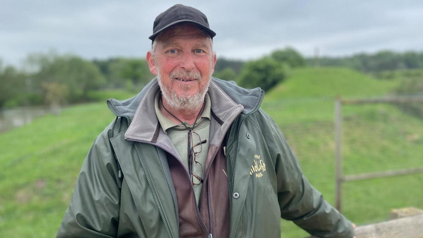 Man wearing waterproof green jacket and hat rests against a fence.