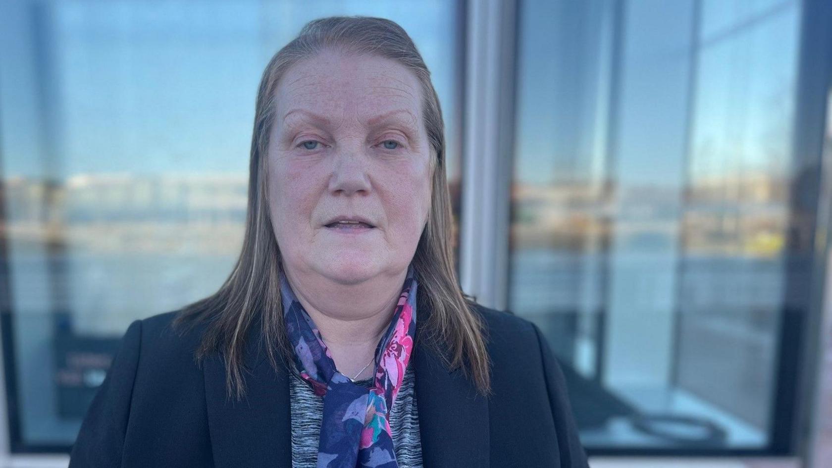 Linda Watson stands in front of windows reflecting a blurry but bright landscape. She has straight hair to her shoulders and is wearing a navy jacket and a blue, lilac and pink flowery scarf over a silver necklace.