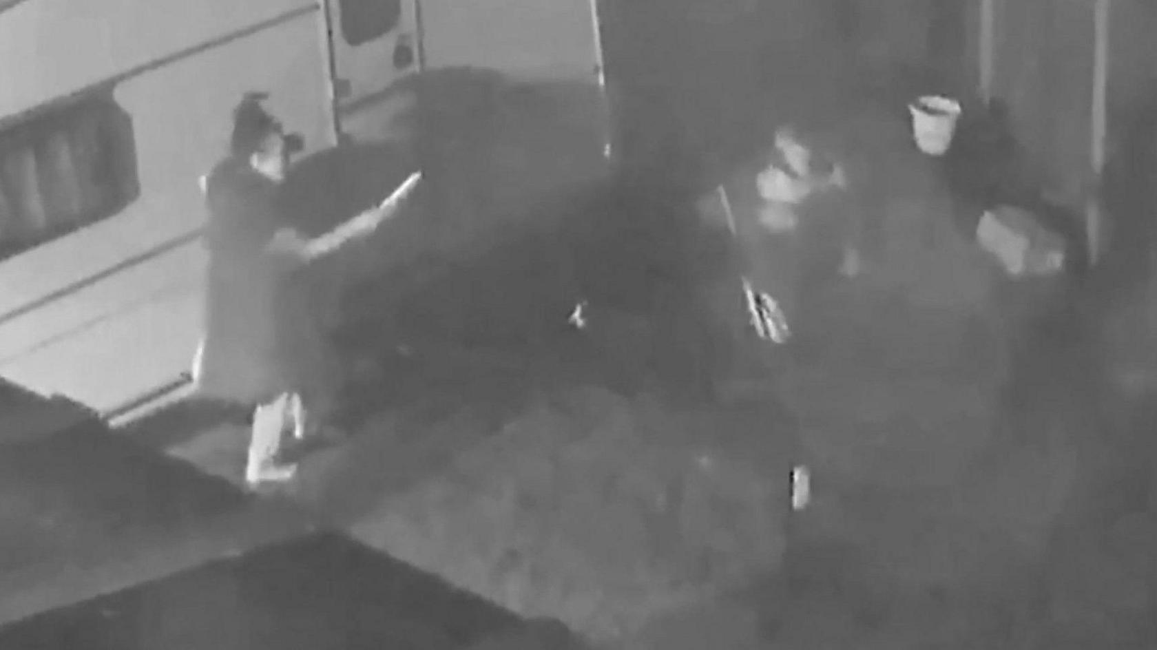 A black and white CCTV image of a woman on the left, next to a caravan, standing in a garden while pointing a knife at a man who appears to be sitting down. 