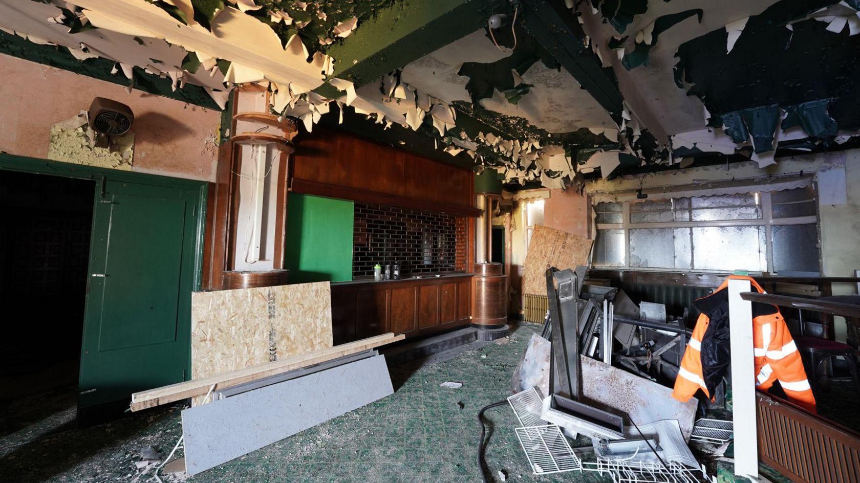 A former pub which has a wooden bar and green tiled flooring. Wallpaper is hanging down from the roof and there are metal sheets over the windows. An orange high vis jacket is hanging inside.