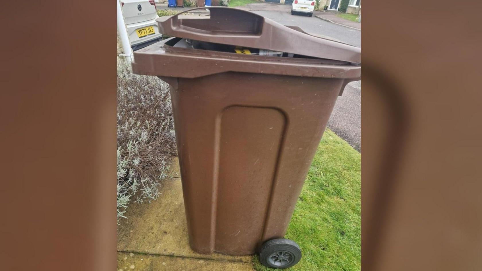 A full brown bin on some paving near a patch of grass 