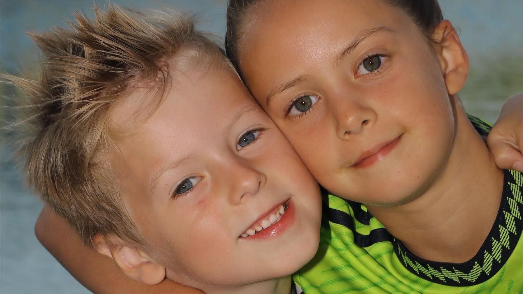 Two children hug and smile at the camera. The boy on the left has short blonde hair and the girl has long brown hair.