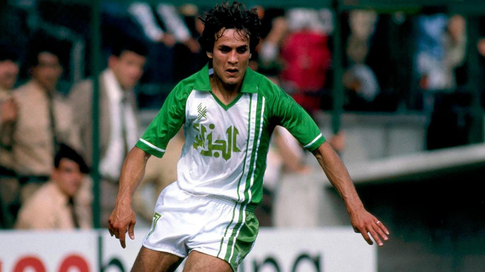 Algeria's Rabeh Madjer seen in action at the 1982 World Cup in Spain. He is wearing predominantly white shorts with green vertical trim on one side and a matching shirt which sees the same green lines carry on up from the shorts onto Madjer's torso to meet a large green collar and green sleeves. Written on the chest, also in green, is the name of Algeria in Arabic script. Madjer is looking down at a ball which is out of shot and leaning to his left, with arms slightly spread for balance, as he looks to turn. His dark hair is bouncing slightly as he moves. Blurred in the background are members of the crowd, including what appears to be some members of stadium security dressed in khaki colours