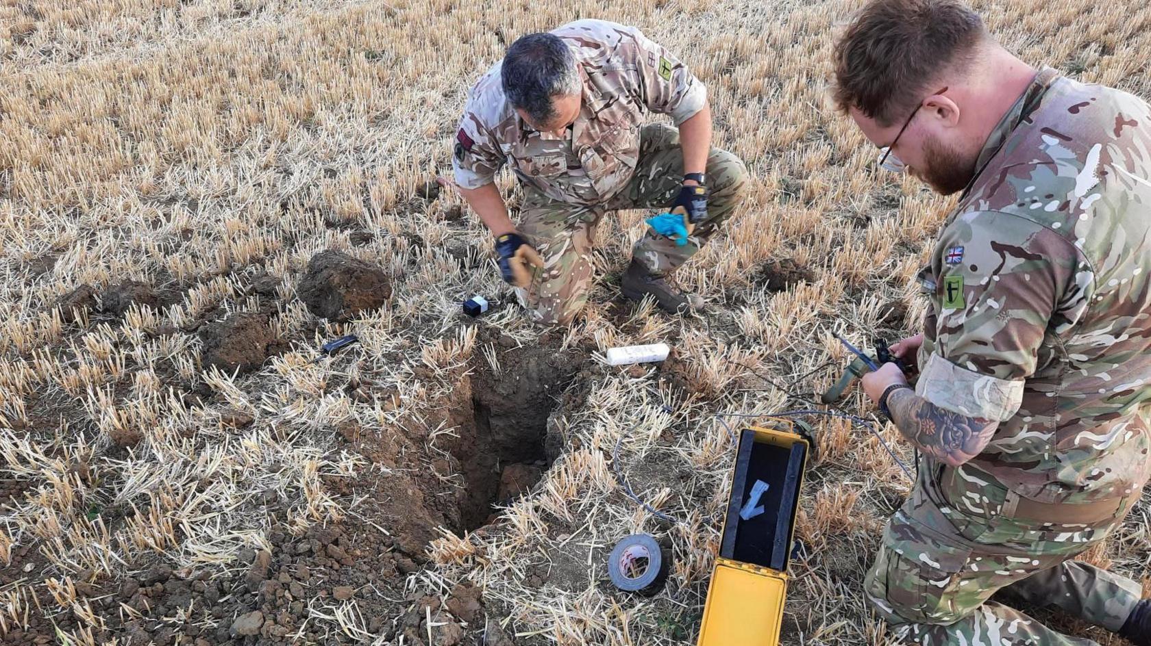 Two men in camouflage Army uniform are looking at a bomb in a pit in a field.
The men have some kind of yellow and black monitoring device. The field is brown and stubbly