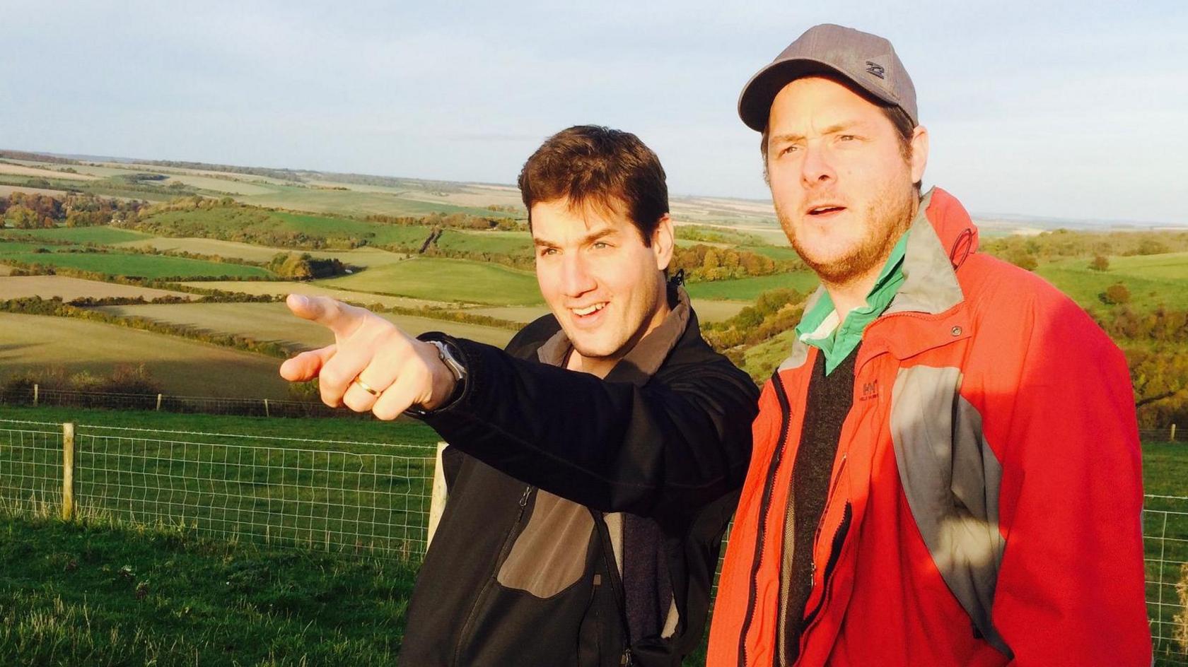 Dr Tom Pelly and his brother. Dr Pelly is pointing at something in the distance. They are in a remote location, with green hills and fields behind them.