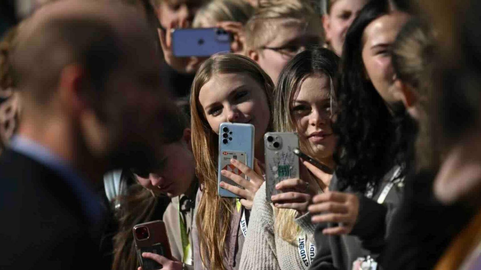 A crowd of young people pointing their cameras to a blurred image of a man in a dark jacket and blue shirt.