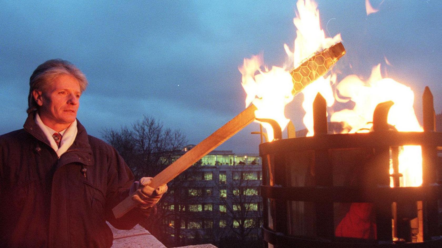 Bruno Peek is wearing a coat with a white scarf. He is holding a long wooden torch to light a beacon on top of St James's church tower in London.