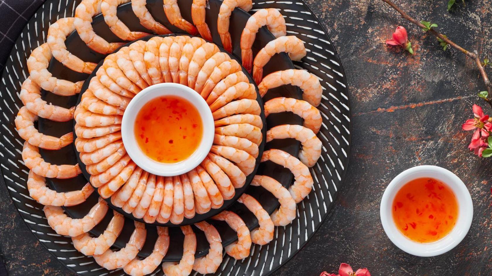 A plate full of prawns laid out on a plate in ring formation. To the right of the plate is an orange sauce