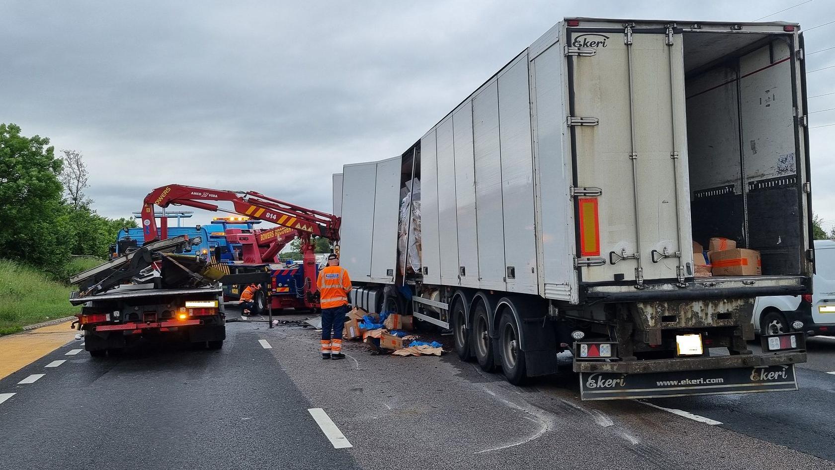 A white large lorry has boxes spilling out the side, a crane is to the left-hand side and a man stands near the area in a high-vis jacket