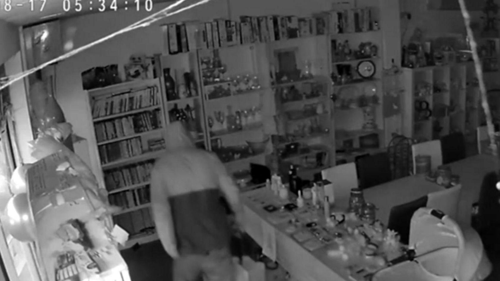 Black and white still of CCTV footage showing a person in a white and grey coat walking around a charity shop carrying a large bag