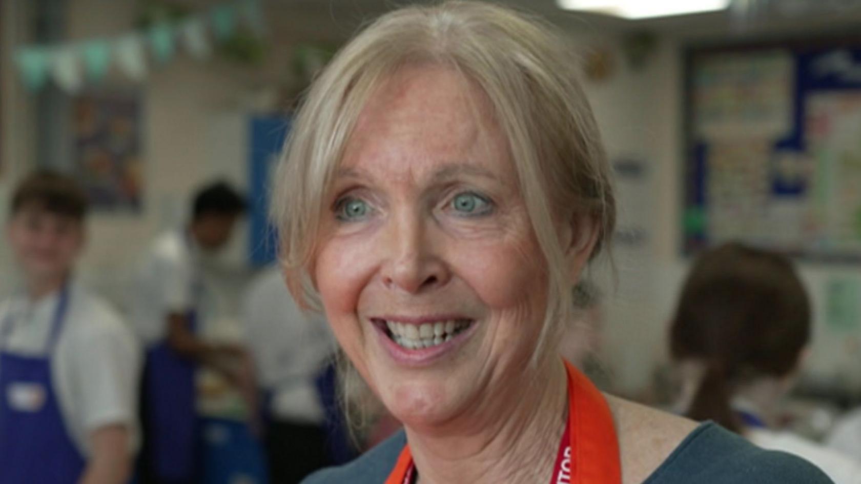 Kate Percy at a school. She is smiling into the camera, while pupils cook behind her.