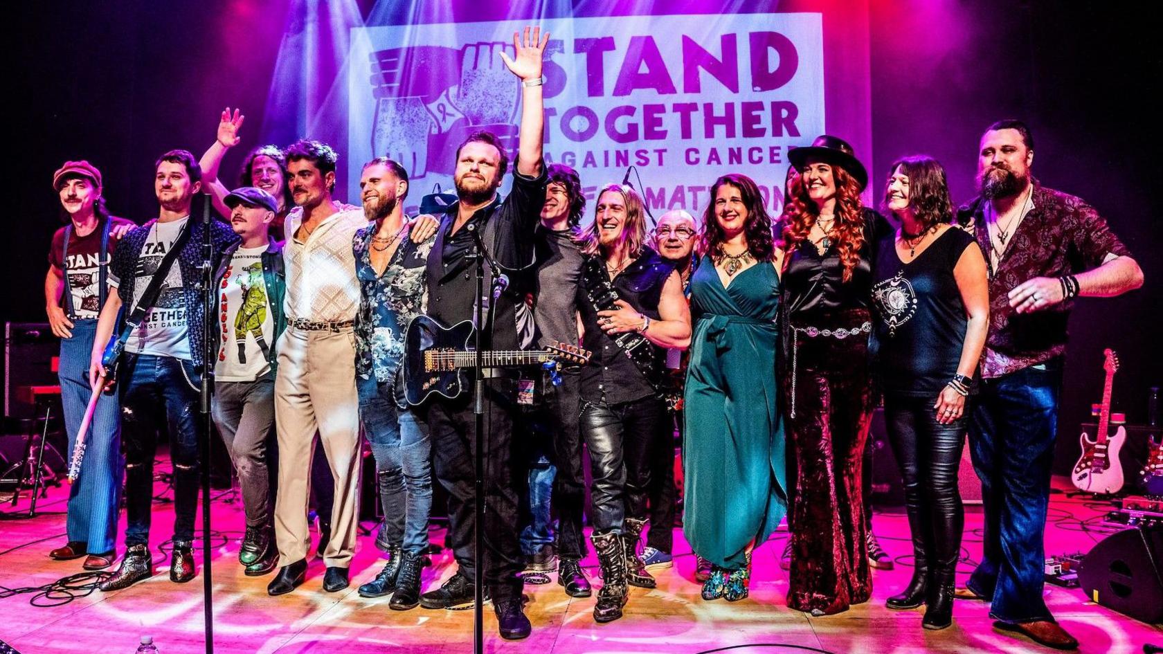 Fourteen musicians gathered together in the centre of a stage lit with pink lights to hank thank the crowd at the end of a show
