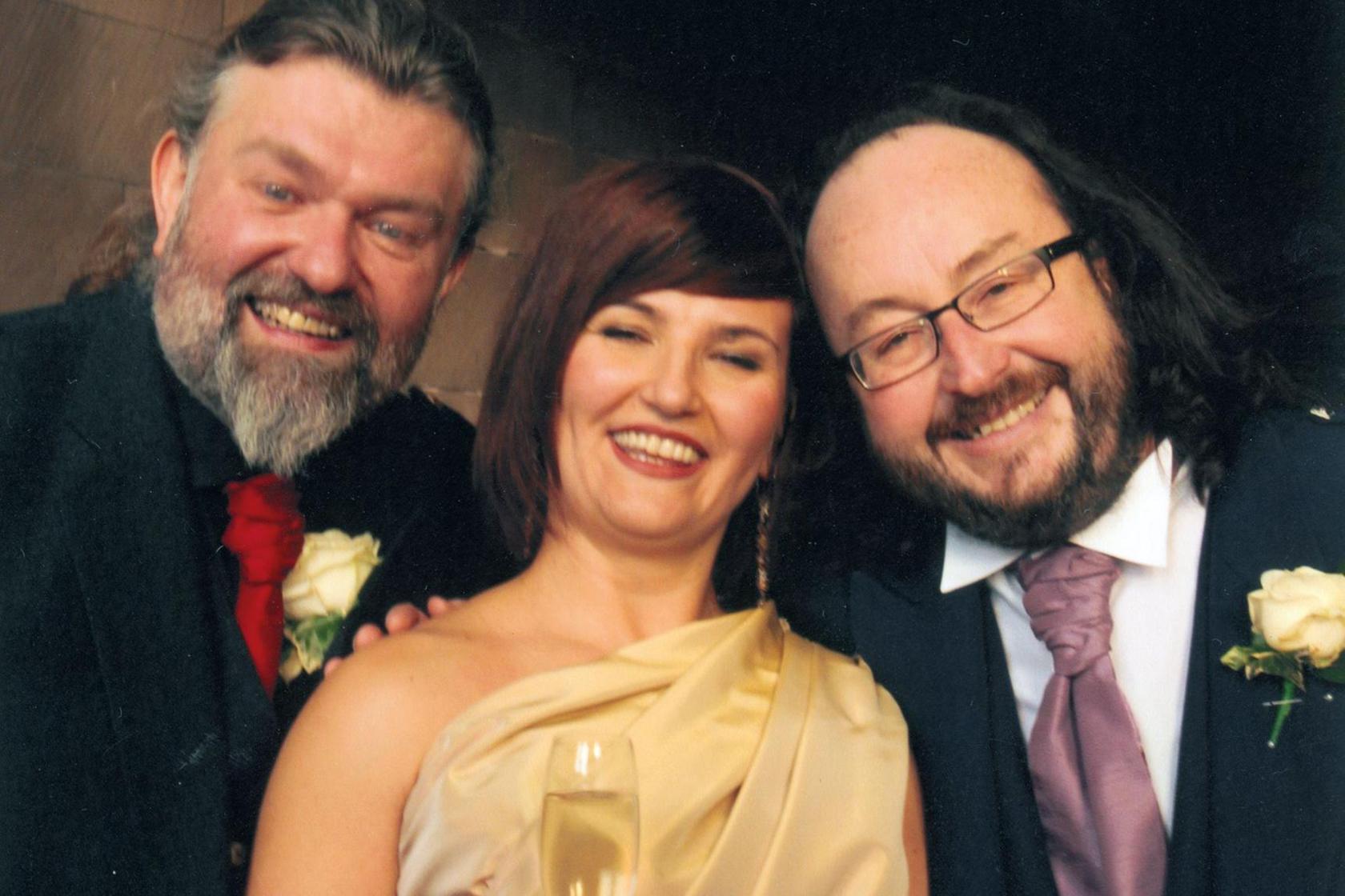 A picture taken at the wedding of Lili and Dave. Lili is in the middle of the shot and is wearing a cream wedding dress that comes off the shoulder. She is carrying a bouquet of orange and cream flowers and is carrying a glass of champagne in her hand. On her right is her husband, Dave. He is looking overjoyed and is smiling at the camera and wearing a purple tie, smart jacket and kilt. Si is on the left of the shot. He is wearing a red tie, smart jacket and a kilt and has a big smile on his face.