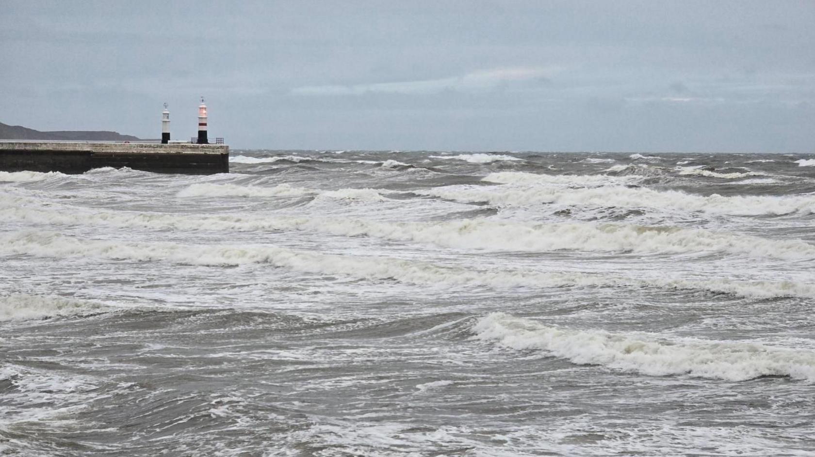 Choppy grey and white waves crash into the shore. An illuminated red light shines from the top of a small lighthouse at the end of a pier.