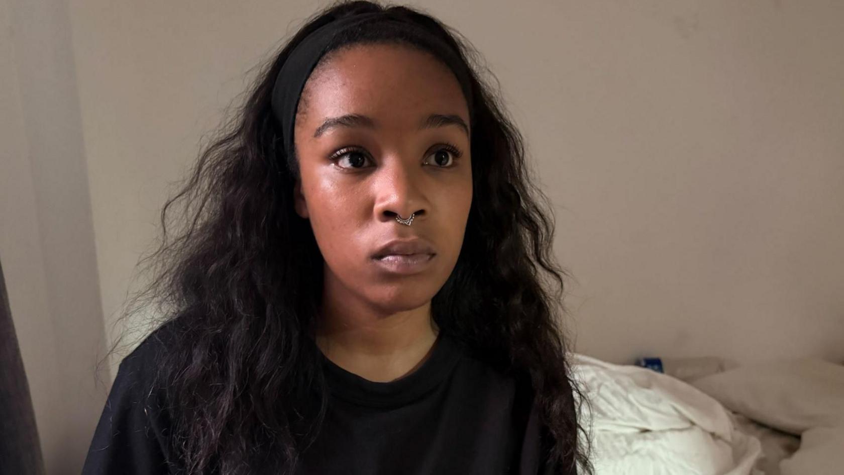 Isabel Rose is seen wearing a black top and a small nose ring, sits facing the camera indoors.