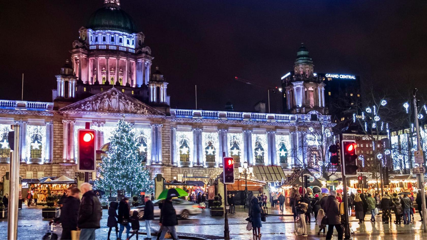 Belfast City Hall lit up with bright white stars in every alcove. Stalls from a Christmas market are visible in front, and there is a big Christmas tree lit up with white lights at the front centre of City Hall. There are a lot of people walking along the street.