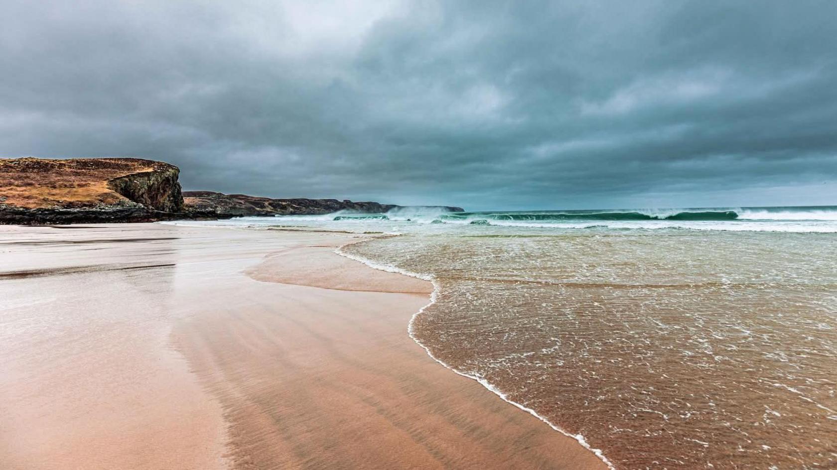 A beach on Lewis. The sky is a mixture of dark blue and grey. There are dark cliffs in the background. The beach in the foreground is light coloured. The water is jade coloured. There is a shallow tide coming in. Some of the waves are crashing on to the beach.