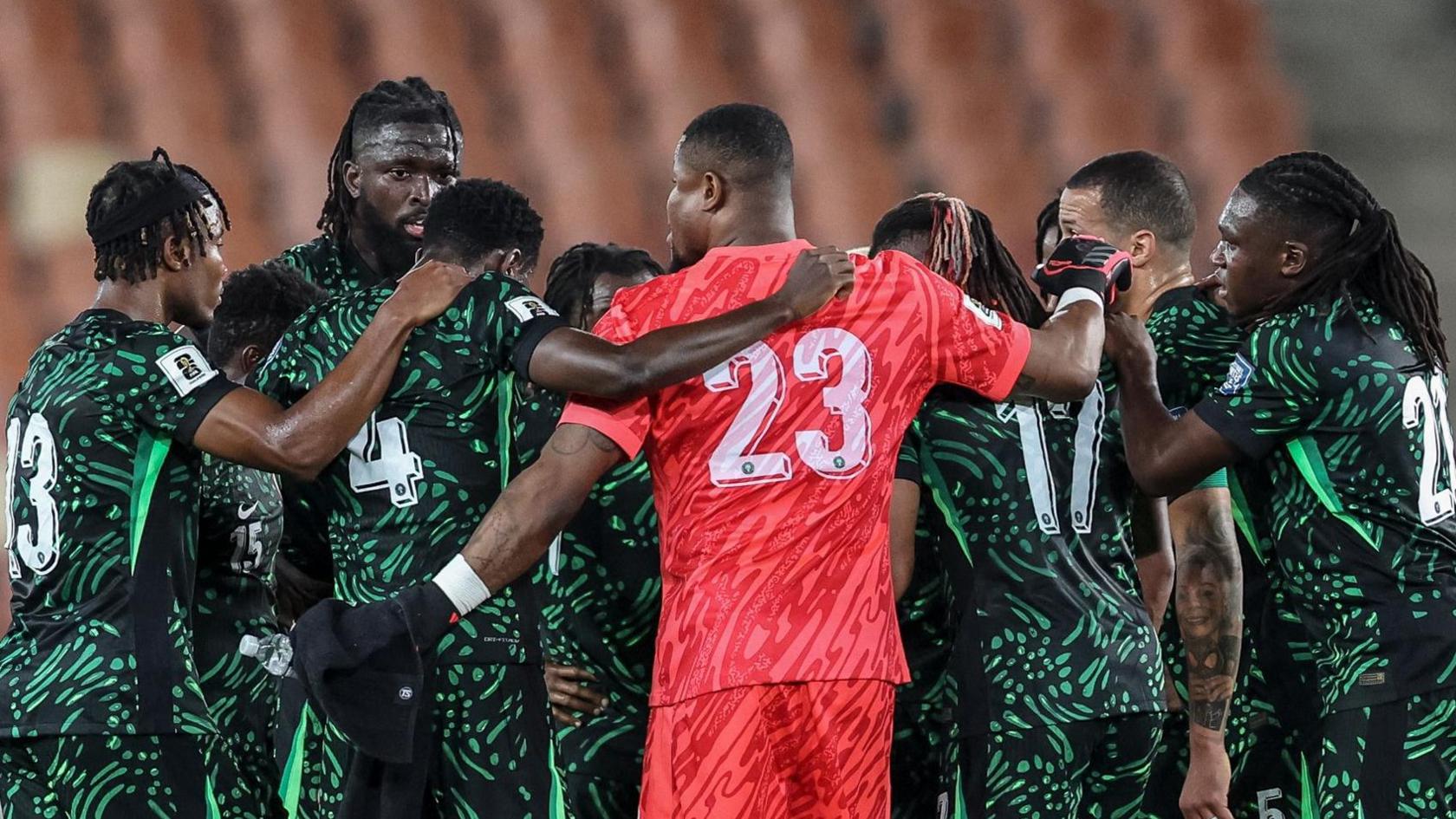 Nigeria players form a team huddle during a match with arms draped around shoulders. All but one are wearing green and black kit, except for goalkeeper Stanley Nwabali who is in the centre of the shot with his back to the camera wearing pink kit with a white number 23