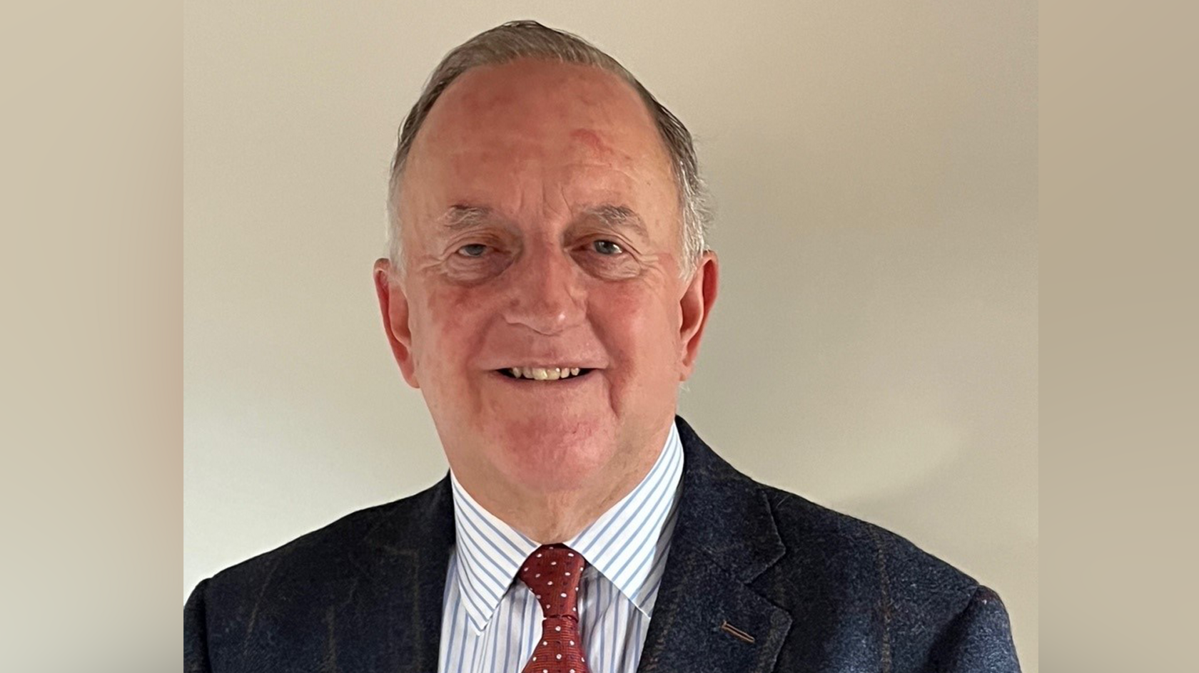 Stuart Clancy smiling. He is wearing a navy jacket with a stripy shirt underneath and a red spotty tie. He has short grey hair.