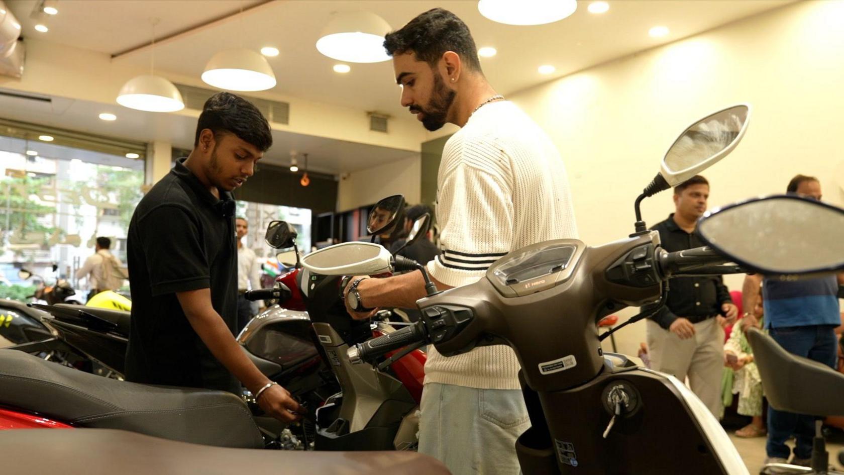 Two men - one in a black t-shirt and the other in white - looking at two-wheelers inside a showroom in Mumbai.
