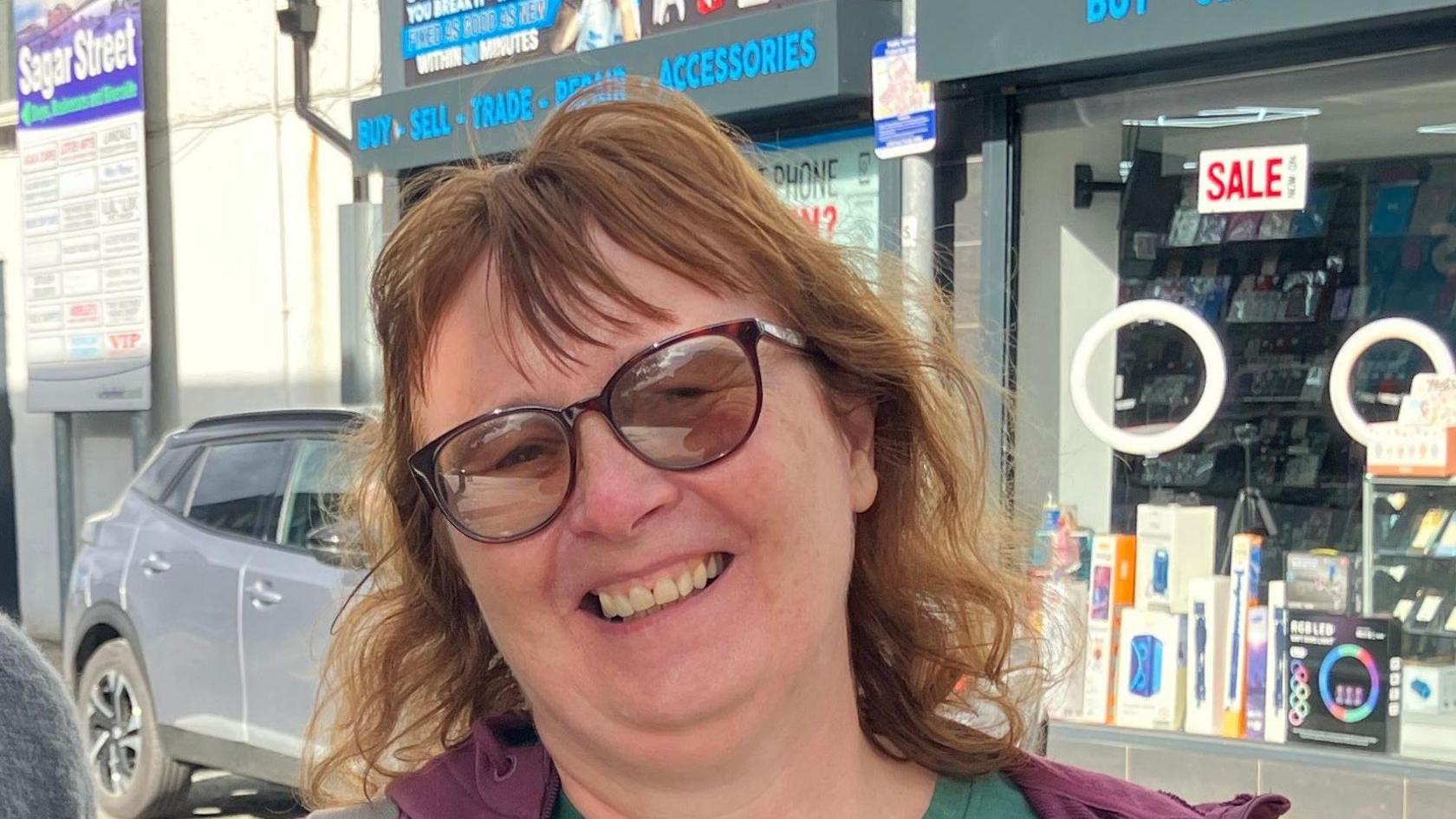 A woman with wavy brown hair and sunglasses smiles at the camera. She is wearing a purple hoodie and standing on a street.