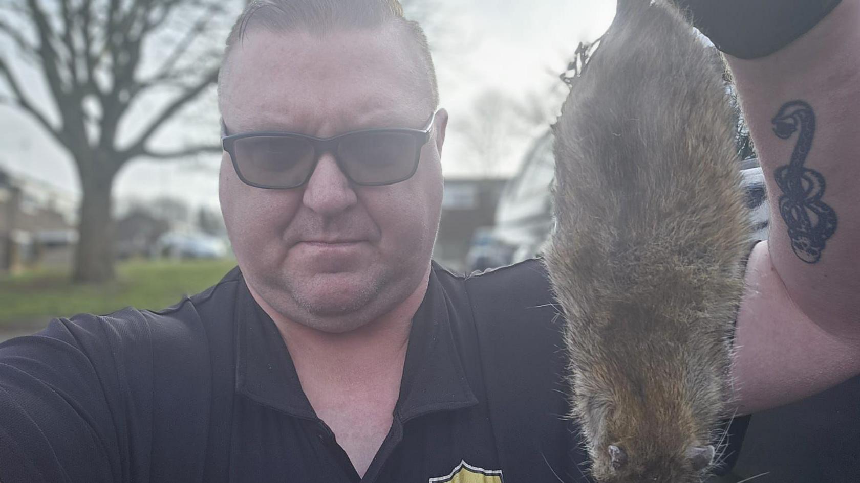 William Timms is wearing sunglasses and a navy blue polo shirt. He has gloves on a tattoo on his forearm. He is holding a dead rat in his left hand.