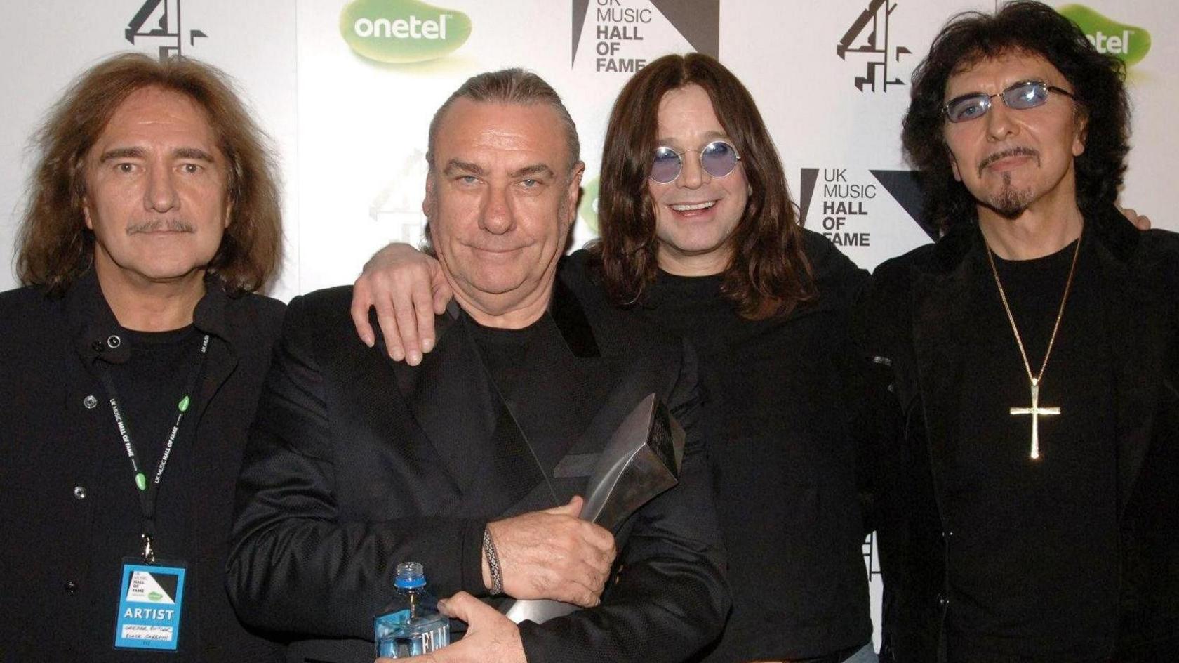 Four men dressed in black standing in front of a backdrop advertising UK Music Hall of Fame, sponsored by Onetel and Channel 4. From left to right are Black Sabbath bassist Geezer Butler, drummer Bill Ward, singer Ozzy Osbourne and guitarist Tony Iommi.