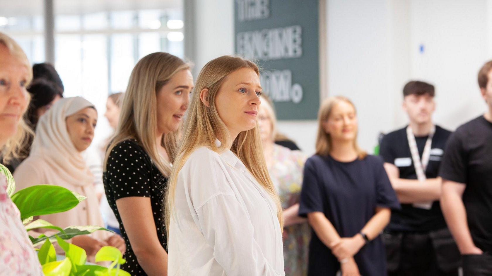 A group of staff members at Deloitte in Bristol listen to a presentation. Two women are at the centre of the picture, both blonde. One is wearing a white shirt and the other is wearing a black short-sleeved dress with white spots on it