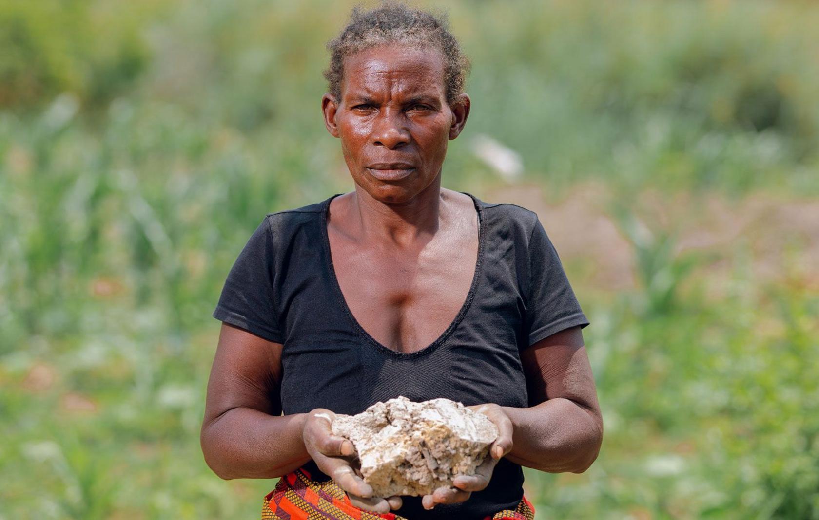 A photo portrait of Mary, a Zambian farmer. She stands in the centre of the frame looking pensive and holding up a large lump of white contaminated crop soil, with both hands. There is greenery behind her and she wear a black top and patterned skirt. 
