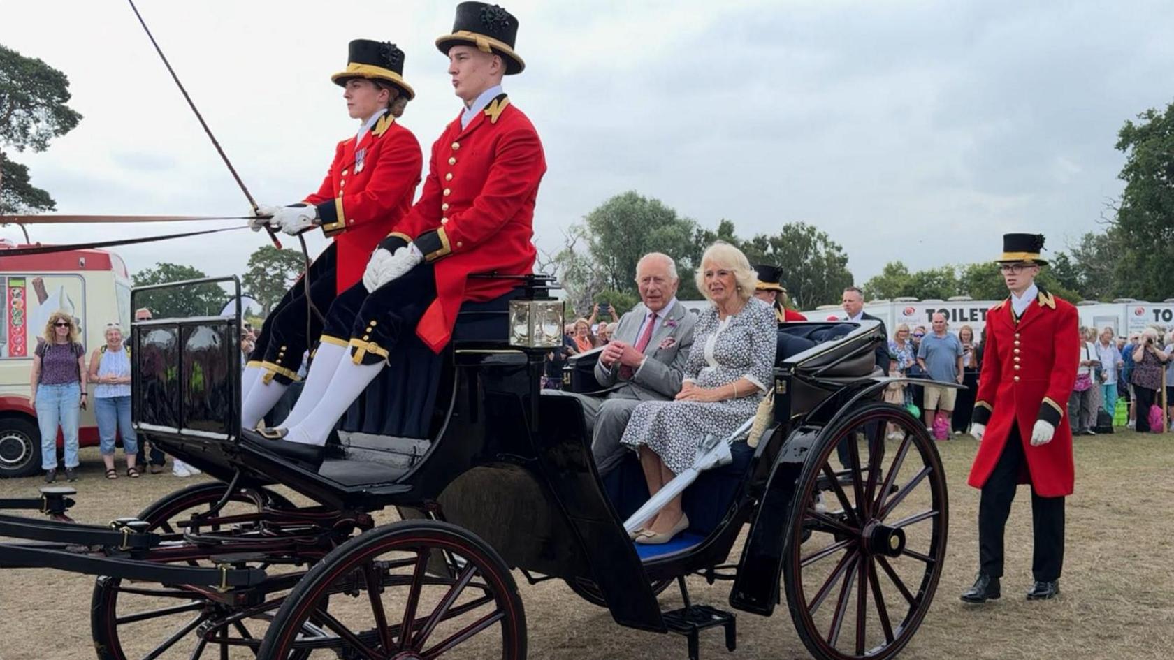 King Charles III and Queen Camilla riding in the back of a horse-drawn carriage to get around the Sandringham Flower Show. King Charles III who is sitting on the left hand side of the carriage is wearing a pale grey suit with a burgundy tie and sitting next to him is Queen Camilla, who is wearing a black and white floral dress. There is a man and woman sat at the top of the carriage and another man following behind them.