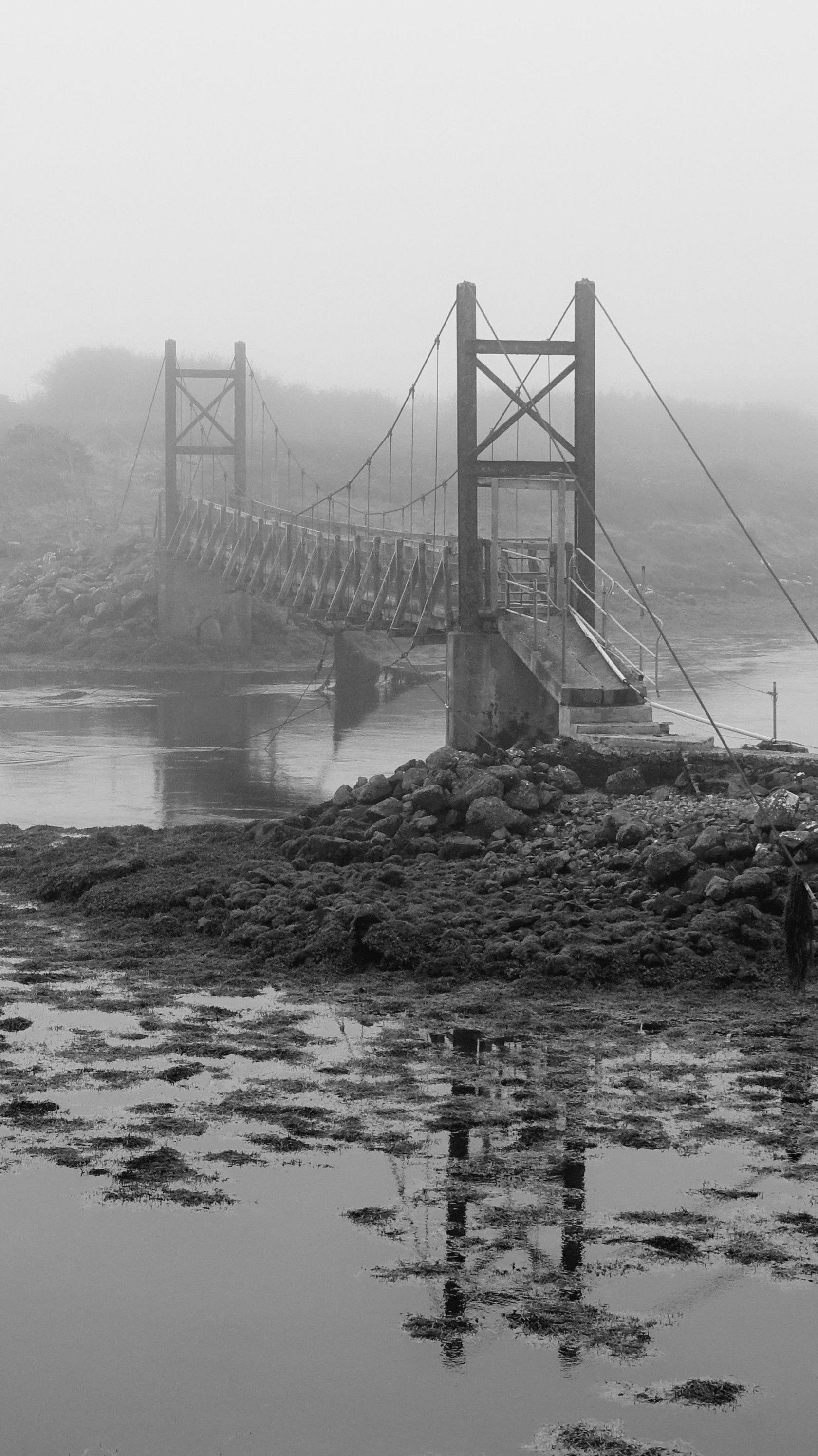 Black and white image of a bridge with a large pile of rocks and boulders at either end and seaweed and debris floating around the rocks in the water below - the sky is grey and overcast 