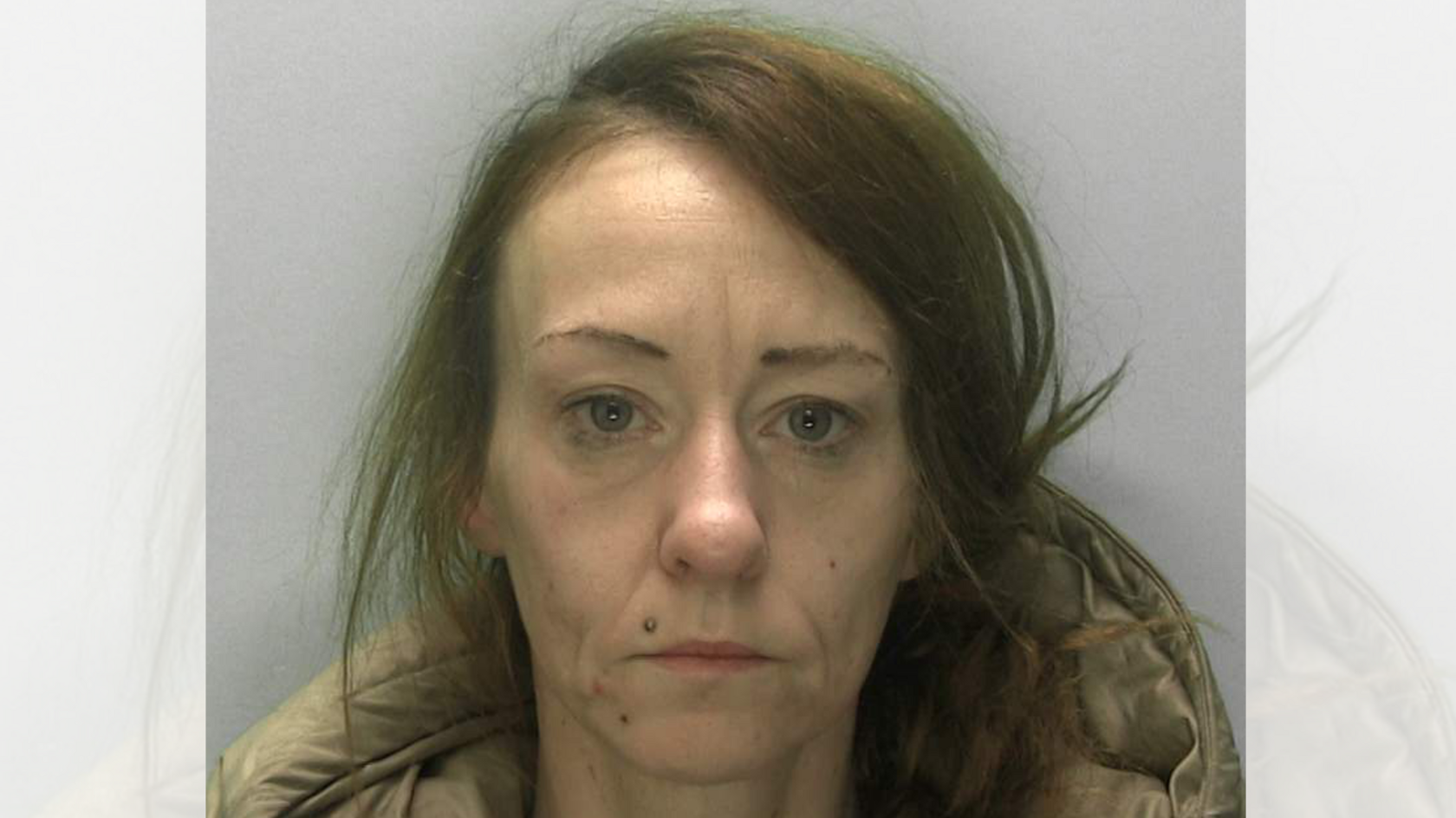 A middle-aged woman looks straight at the camera with her brown hair parted to the right. She has a mole on her upper lip and is wearing a light-brown overcoat, of which only the hood is visible.