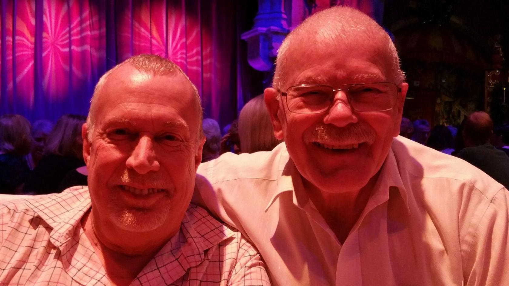 Stuart and Peter wearing shirts at an evening event indoors. They are smiling and lit up by pink and purple lighting.