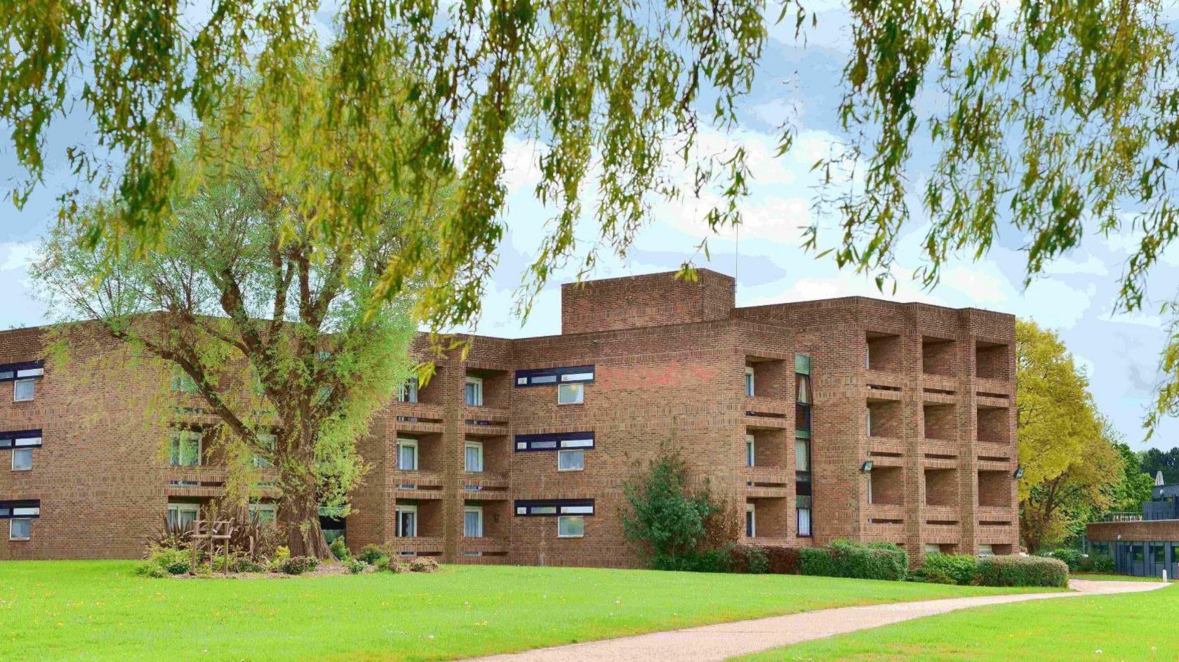 Brick building with 28 visible windows across three stories. There is a pavement passing through a grassed areas and a large tree.