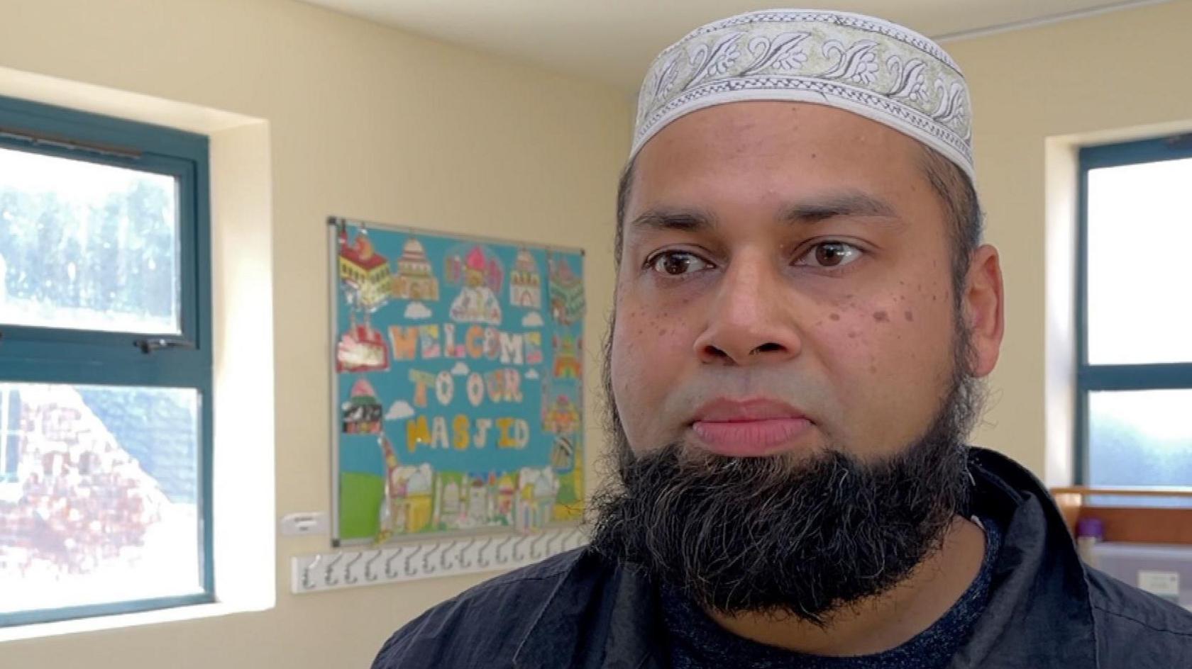 Bulbul Miah stands inside the Jame Masjid mosque. He is wearing a skullcap and has a black beard. In the background, a colourful sign reads "welcome to our masjid".