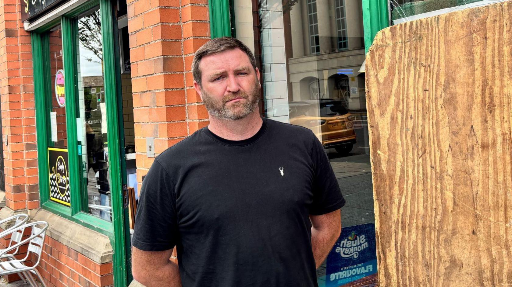 Gareth McKinstry, with short dark hair and a black t-shirt, standing in front of his chipshop