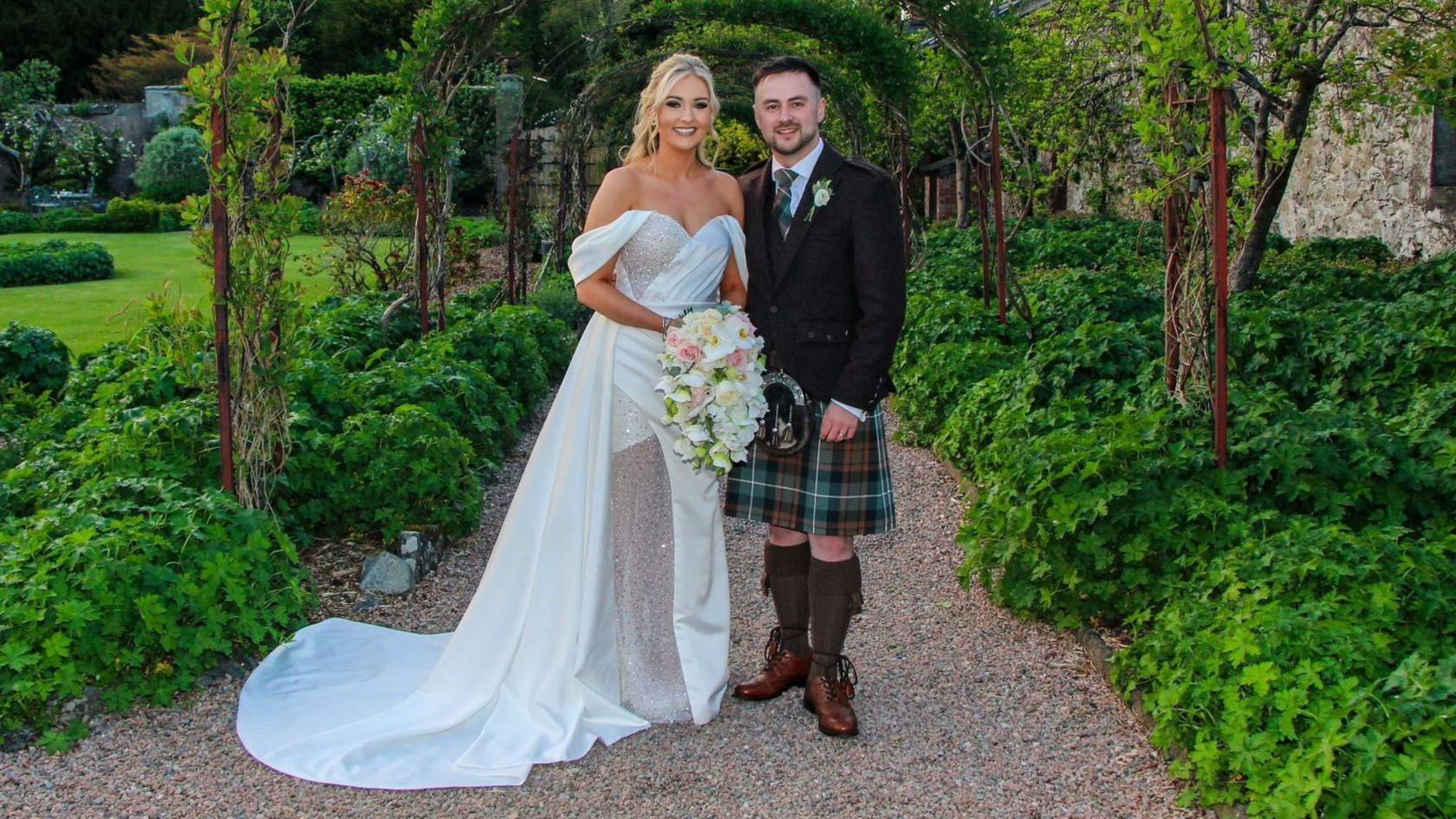 The bride is wearing a white dress with off the shoulder sleeves and glitter details on the bodice and leg. She is standing on the left. The groom is wearing a kilt and suit jacket. They are standing beneath a garden arch and smiling at the camera.