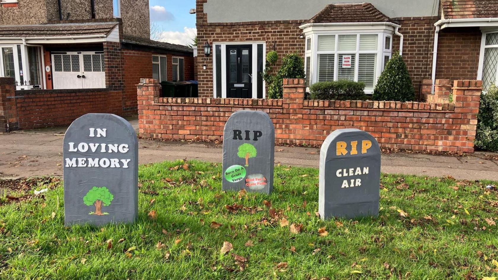 Three signs made to look like gravestones are on a grass lawn. The signs read 'In Loving Memory' 'RIP' and 'Clean Air' alongside drawings of trees.