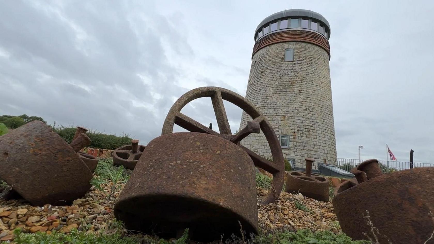 A tall stone windmill with a glass top. In front of it on the ground are rusty pieces of industrial metal.