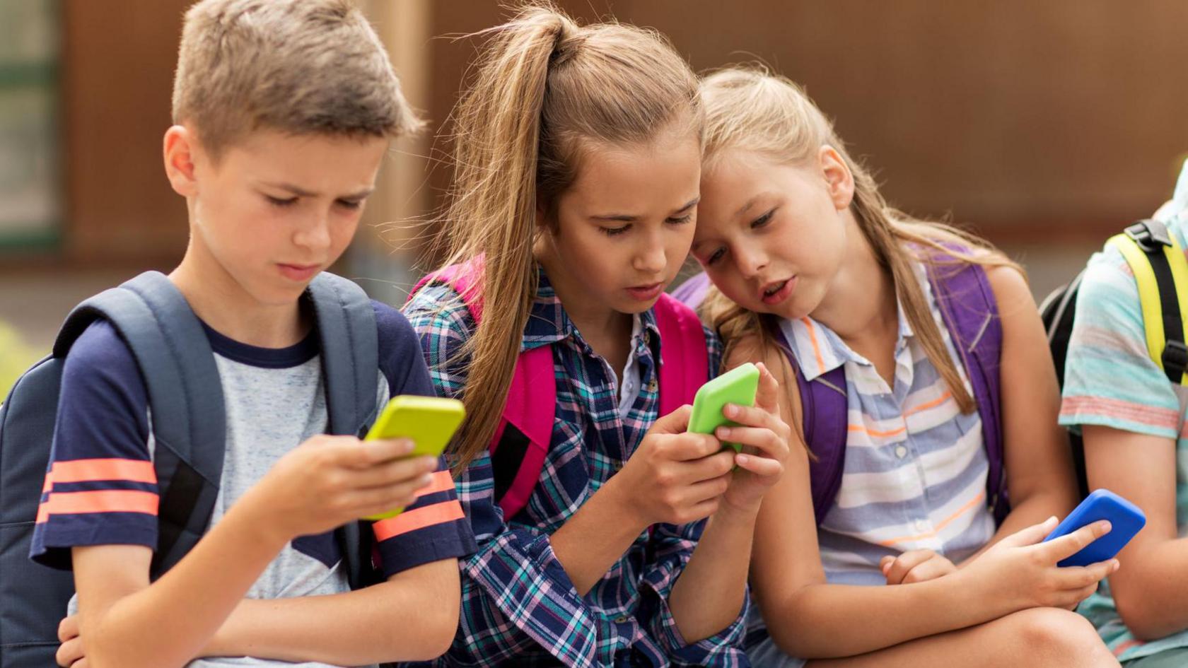 Three children, each holding mobile phones, sitting beside each other on a wall. To the left is a boy with short brown hair, wearing a grey t-shirt and holding a yellow-backed phone. Two girls with shoulder length brown hair holding green and blue backed phoned are beside him.