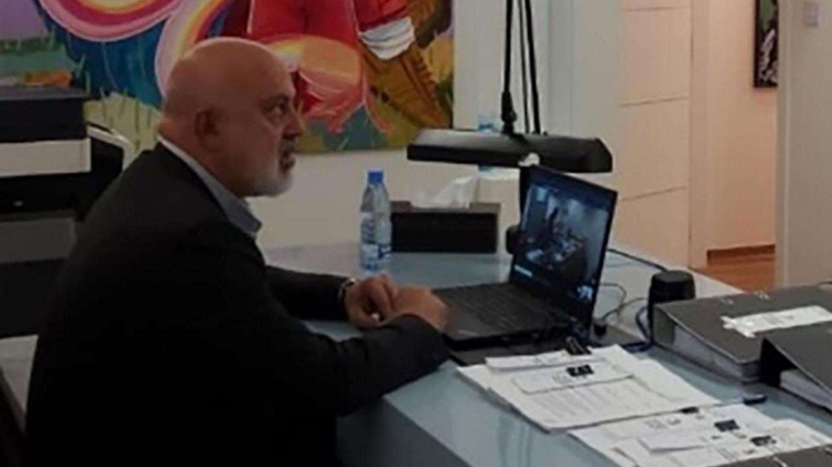 Nazem Ahmed sits at a desk with a laptop and paperwork in front of him. He is wearing a suit. He is bald.